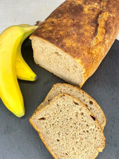 Dinkel-Bananenbrot Puristo Bananenbrot mit goldbrauner Kruste, lockerer Krume, aufgeschnitten neben reifen Bananen.
